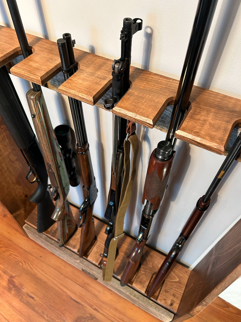 May include: A wooden gun rack displaying several rifles and shotguns. The rack is constructed from stained wood and features slots designed to securely hold the firearms. Various rifles and shotguns are visible, showcasing diverse designs and finishes. The rack is mounted on a wall.