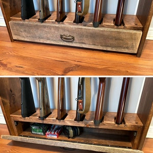 May include: A wooden gun rack with slots for several rifles. The rack features a drawer with a metal handle. The rifles are in various colours, including black, brown, and wood grain. The rack is made of stained wood.