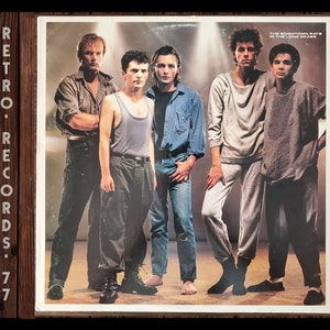 May include: A vintage vinyl record album cover featuring a band of five men. The album cover is titled "The Boomtown Rats In The Long Grass". The band members are standing in a row, with the lead singer in the center. The album cover is a retro style with a faded color scheme.