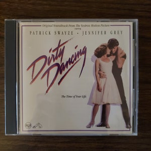 May include: A CD case for the soundtrack to the movie "Dirty Dancing" starring Patrick Swayze and Jennifer Grey. The case features a photo of the two actors dancing, with the movie title in large, purple letters. The text "The Time of Your Life" is printed below the title.