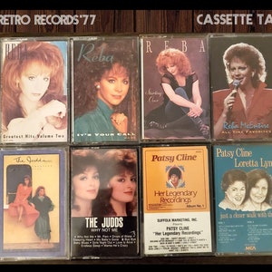 May include: A collection of eight vintage cassette tapes featuring country music artists Reba McEntire, The Judds, and Patsy Cline. The tapes are displayed on a wooden surface.