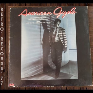May include: A black and white vinyl record cover for the soundtrack to the film "American Gigolo". The cover features a man in a suit standing in a room with a window behind him. The text on the cover reads "American Gigolo", "Original Soundtrack Recording", "Music Composed by Giorgio Moroder", "Blond/"Call Me"", "Cheryl Barnes/"Love & Passion"", and "Told Her".