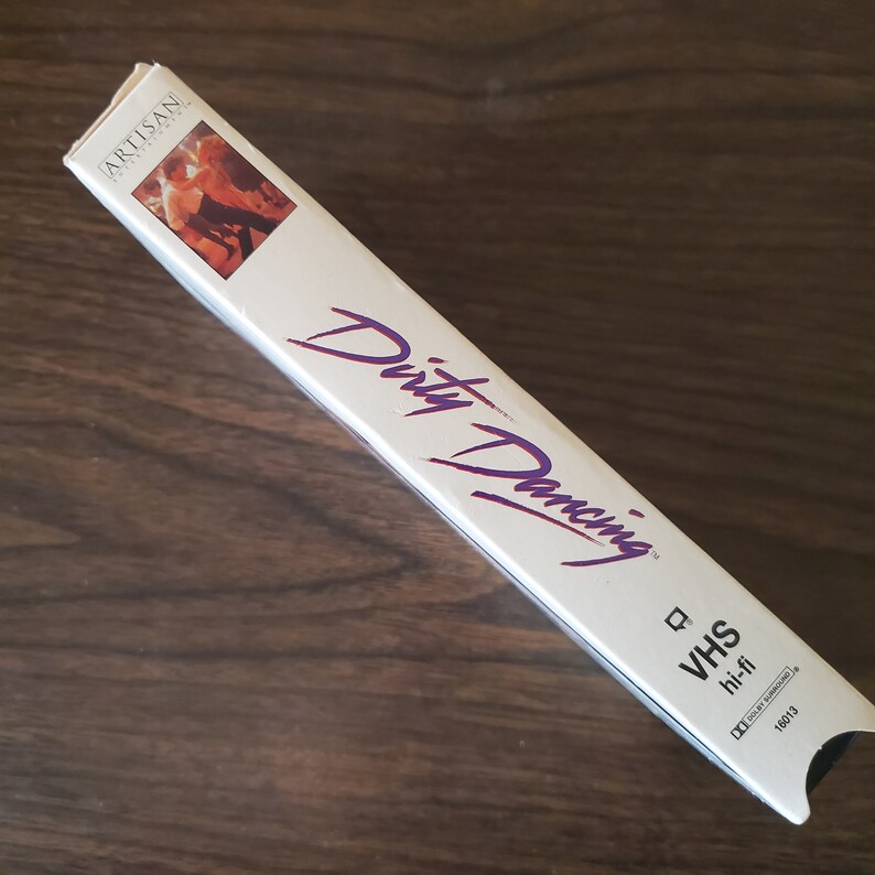 May include: A white VHS tape case with the title "Dirty Dancing" in purple lettering. The Artisan Home Video logo is in the top left corner. The case also says "VHS hi-fi" and has a barcode.