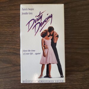 May include: A VHS tape cover for the movie "Dirty Dancing" featuring Patrick Swayze and Jennifer Grey. The cover has a white background with the movie title in purple and pink lettering. The tagline "Have the time of your life...again!" is printed below the title. The cover also features a photo of Swayze and Grey dancing.
