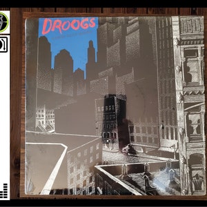 May include: A black and white album cover with the title "Droogs" and the text "Stone Cold Groove" in white. The cover features a black and white illustration of a city skyline with buildings and rooftops.