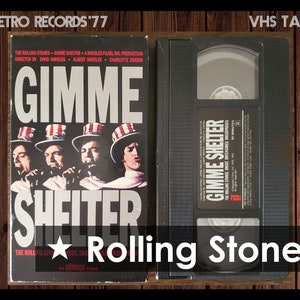 May include: VHS tape case with a black cover and a label that reads "Gimme Shelter" and "The Rolling Stones Uncut Uncensored Unsurpassed". The label also includes the title "Gimme Shelter" in large white letters. The case is on a wooden surface.