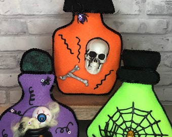 May include: Three felt Halloween potion bottles. The first bottle is orange with a skull and crossbones, the second is purple with a cauldron and an eyeball, and the third is green with a spider web and a spider.