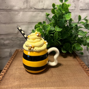 May include: A yellow and black striped ceramic mug with a white handle. The mug is topped with a yellow whipped cream-like substance and decorated with small bee figurines.