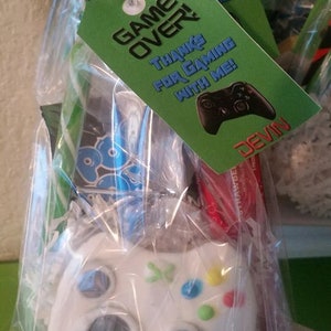 May include: A green and white favor bag with a green tag that says "Game Over! Thanks for gaming with me! Devin". The bag contains a white plastic video game controller with colorful buttons.