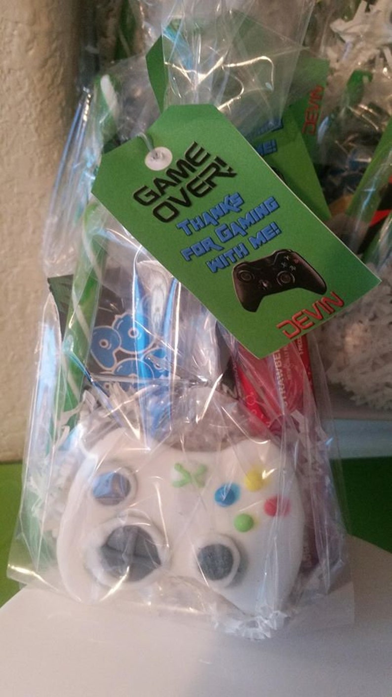 May include: A green and white favor bag with a green tag that says "Game Over! Thanks for gaming with me! Devin". The bag contains a white plastic video game controller with colorful buttons.