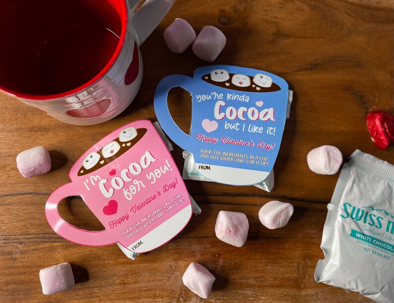 May include: Two Valentine's Day cocoa-themed gift cards shaped like mugs, one pink and one blue, with the phrases "I'm Cocoa for you!" and "You're kinda Cocoa but I like it!" printed on them. The cards are accompanied by marshmallows and a packet of Swiss Miss hot drink mix.