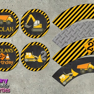 May include: Construction themed cupcake wrappers and toppers with yellow and black stripes, featuring images of construction vehicles and a sign with the number 2 and a traffic cone. The toppers also include the text "Nolan's 2nd Birthday".