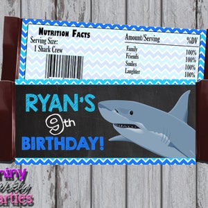 May include: A blue and white chevron patterned chocolate bar wrapper with a shark illustration and the text "RYAN'S 9th BIRTHDAY!" on a black chalkboard background. The wrapper also includes a nutrition facts label with the text "Serving Size: 1 Shark Crew" and a list of ingredients with percentages.