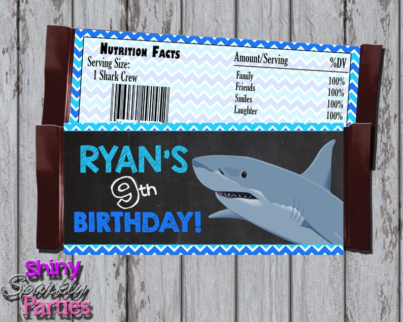 May include: A blue and white chevron patterned chocolate bar wrapper with a shark illustration and the text "RYAN'S 9th BIRTHDAY!" on a black chalkboard background. The wrapper also includes a nutrition facts label with the text "Serving Size: 1 Shark Crew" and a list of ingredients with percentages.