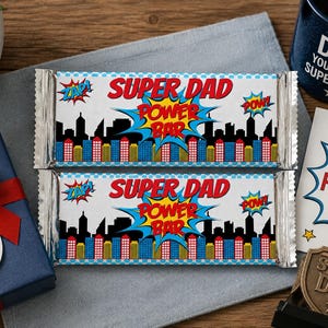 May include: Two "Super Dad Power Bar" candy bars with a comic book style design, featuring a cityscape and the words "Super Dad" in red. A blue mug with "Dad, you're my superhero" and a "Happy Father's Day" card are also present.