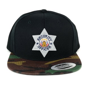 Op de afbeelding: Zwarte snap back pet met camouflageprint en een zilveren stervormige patch met de tekst "Security Officer" in blauwe letters.