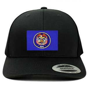 Könnte beinhalten: Schwarze Trucker-Kappe mit einem blauen und goldenen gestickten Aufnäher mit dem Staatssiegel von Utah. Der Aufnäher enthält die Worte "Great Seal of the State of Utah" und "1896". Die Kappe hat eine schwarze Netzrückseite und einen schwarzen Druckknopfverschluss.