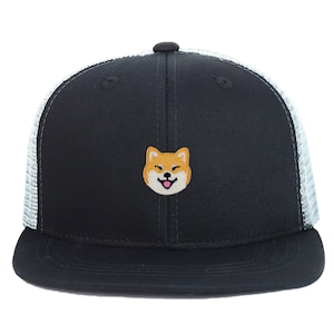 Puede incluir: Una gorra de camionero negra y blanca con una parte trasera de malla. La parte delantera de la gorra presenta un perro Shiba Inu bordado con una expresión feliz.