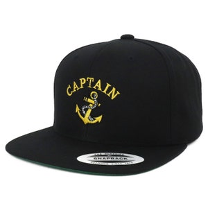 May include: Black and gold baseball cap with the word "CAPTAIN" embroidered in gold on the front. The cap also features a gold anchor with a rope design. The back of the cap has a black label with the text "THE CLASSICS" and "SNAPBACK" in white and "YUPOONG SINCE 1974" in black.