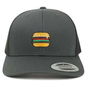 May include: A dark gray trucker hat with a black mesh back. The hat has an embroidered image of a yellow hamburger with red, green, and yellow toppings. The hat has a black sticker on the back that says "The Classics" and "Snapback" with the text "YUPOONG SINCE 1974" underneath.