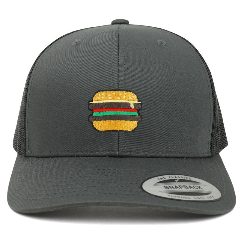 May include: A dark gray trucker hat with a black mesh back. The hat has an embroidered image of a yellow hamburger with red, green, and yellow toppings. The hat has a black sticker on the back that says "The Classics" and "Snapback" with the text "YUPOONG SINCE 1974" underneath.