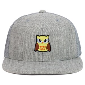 Op de afbeelding: Een grijze en zwarte truckercap met een geborduurde gele uil op de voorkant. De uil heeft oranje ogen en bruine vleugels.
