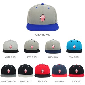 Puede incluir: Una colección de gorras de béisbol con diferentes combinaciones de colores. Las gorras presentan un diseño de cupcake rosa en la parte delantera. Los colores incluyen gris, blanco, negro, azul verdoso, rojo y azul marino.