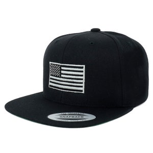 Peut inclure: Une casquette de baseball noire ajustée avec un drapeau américain blanc et noir brodé sur le devant. La casquette a une visière verte et un autocollant blanc qui indique "The Authentic Snapback Yupoong Since 1974".