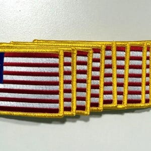 Può includere: Un gruppo di toppe ricamate con il design della bandiera di Betsy Ross. Ogni toppa presenta un cantone blu con stelle bianche, strisce rosse e bianche e un bordo dorato. Le toppe sono rettangolari e sembrano nuove.