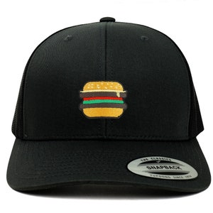 May include: Black trucker hat with a black mesh back. The hat has an embroidered image of a hamburger with yellow cheese, red tomato, green lettuce, and a brown patty. The hat has a black snap closure and a white label that says "The Classics" with a small logo and "Snapback" in a white font. The label also says "Yupoong Since 1974" in a smaller white font.