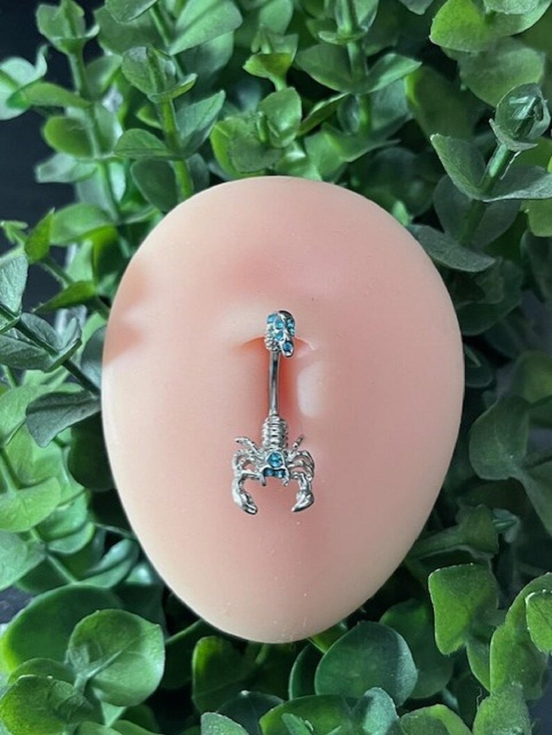 Pode incluir: Um piercing de corpo de prata com um charme de escorpi&atilde;o e pedras preciosas azuis. O escorpi&atilde;o tem uma pedra preciosa azul para o olho.