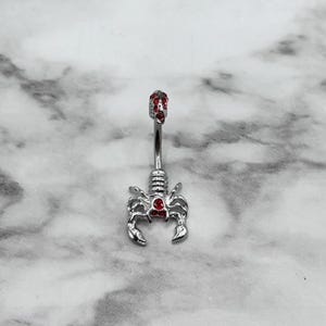 Pode incluir: Anel de umbigo prateado com design de escorpi&atilde;o e detalhes em cristal vermelho.