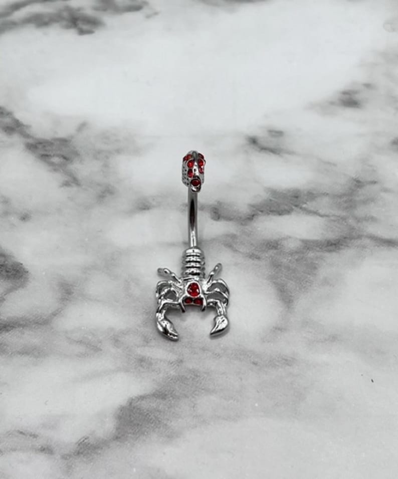 Pode incluir: Anel de umbigo prateado com design de escorpi&atilde;o e detalhes em cristal vermelho.