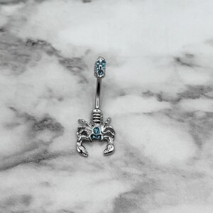 Pode incluir: Um piercing de umbigo de prata com um charme de escorpi&atilde;o e detalhes de cristal azul.