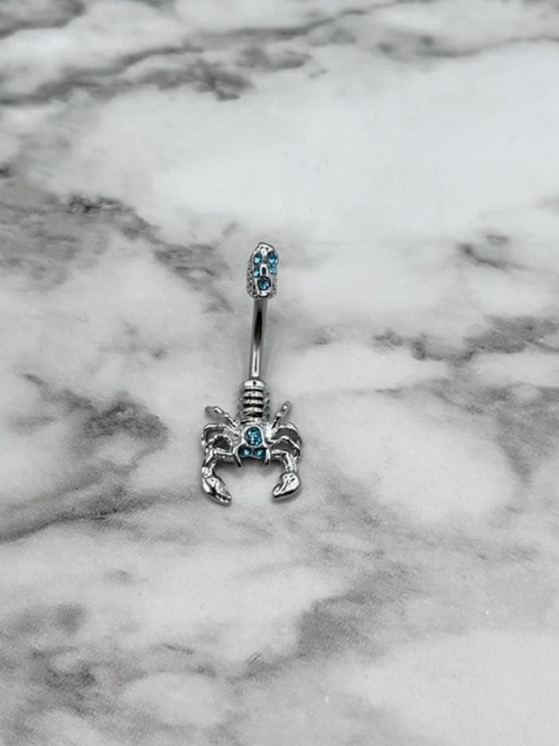 Pode incluir: Um piercing de umbigo de prata com um charme de escorpi&atilde;o e detalhes de cristal azul.