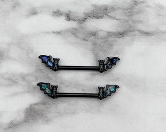 May include: Two black barbell nipple rings with bat charms. The bats have iridescent blue and green wings.