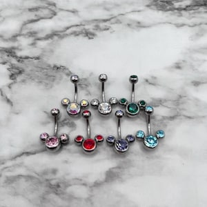 May include: Collection of seven belly button rings with a Mickey Mouse-inspired design. Each ring features a curved barbell with a small ball on top and a cluster of three round, colorful gems at the bottom. Colors include pink, red, purple, and teal.