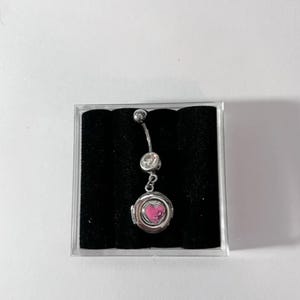 May include: A silver belly button ring with a dangling locket. The locket is round with a pink heart design inside. The ring has two clear, round gems and a small ball at the top.