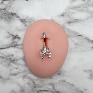 Pode incluir: Piercing corporal de prata com um charme de escorpi&atilde;o. O piercing est&aacute; em um manequim de cor de carne.
