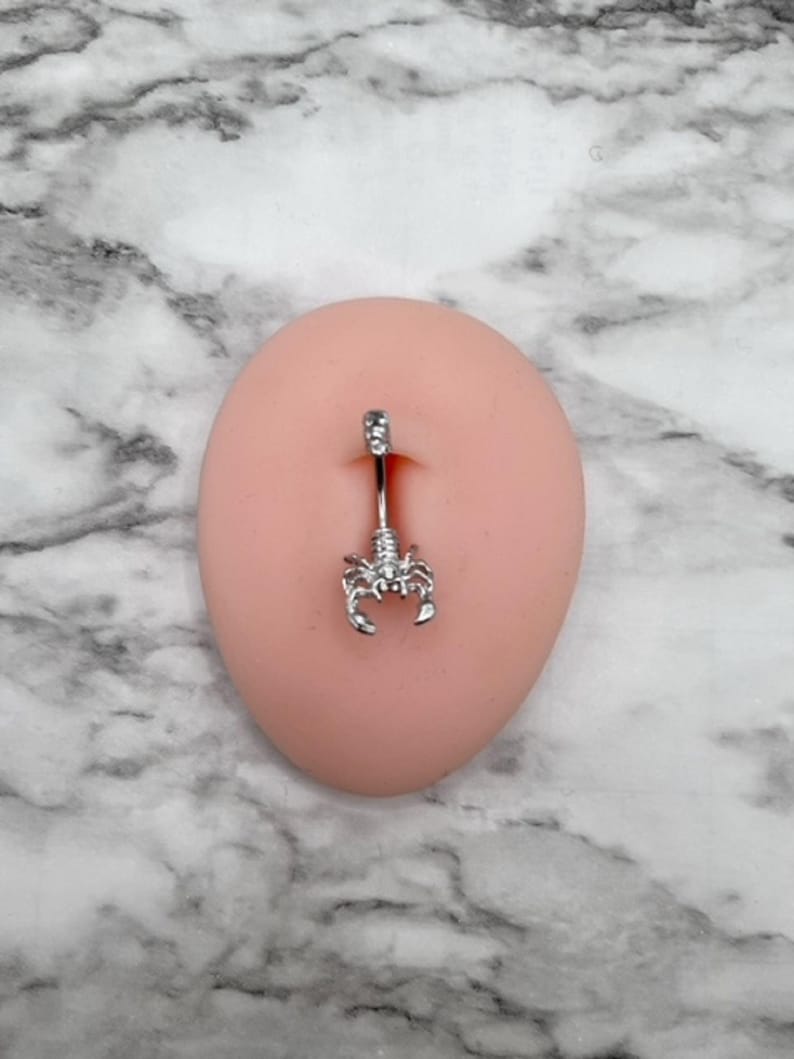 Pode incluir: Piercing corporal de prata com um charme de escorpi&atilde;o. O piercing est&aacute; em um manequim de cor de carne.