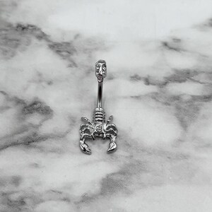 Pode incluir: Piercing corporal de prata com um design de escorpi&atilde;o e pedras transparentes. O escorpi&atilde;o est&aacute; voltado para a frente com a cauda enroscada.