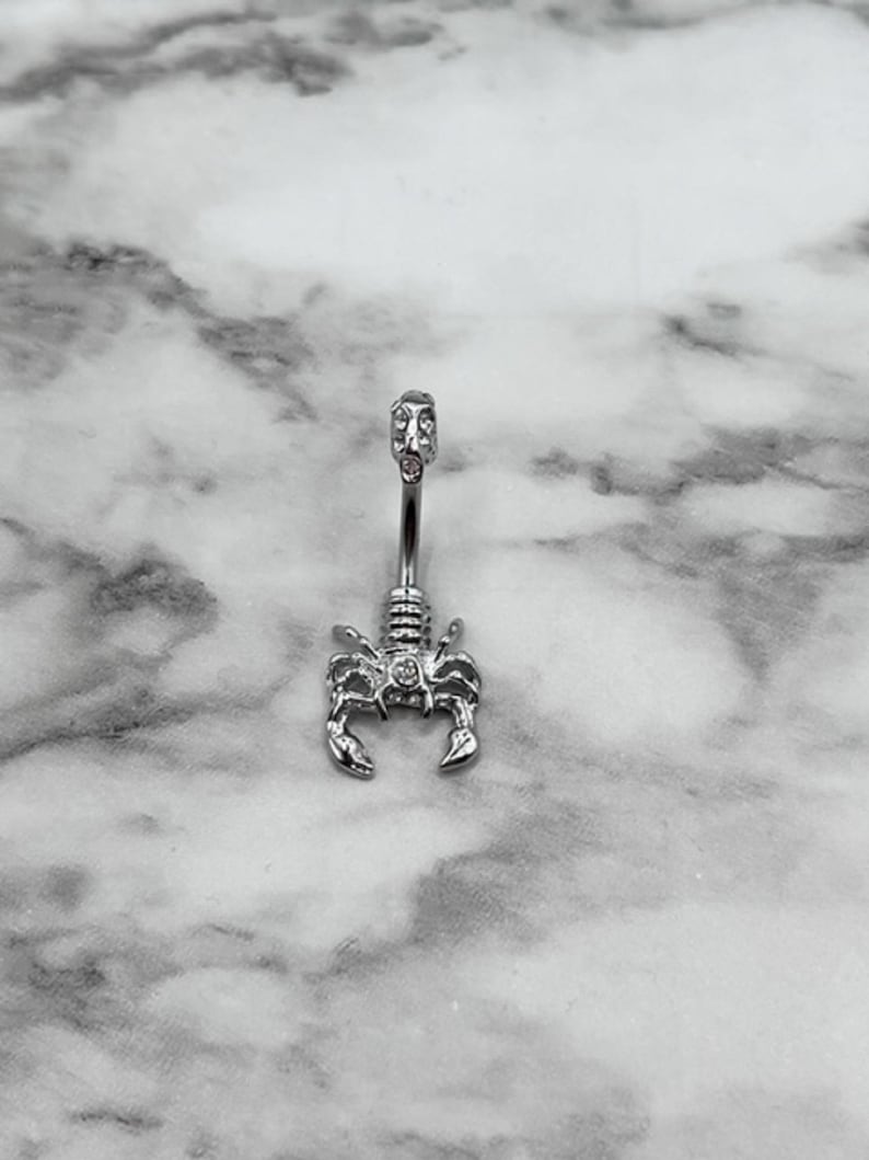 Pode incluir: Piercing corporal de prata com um design de escorpi&atilde;o e pedras transparentes. O escorpi&atilde;o est&aacute; voltado para a frente com a cauda enroscada.
