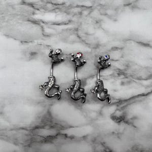 May include: Three silver belly button rings with a lizard design. The lizards have red, blue, and white gemstones for eyes.