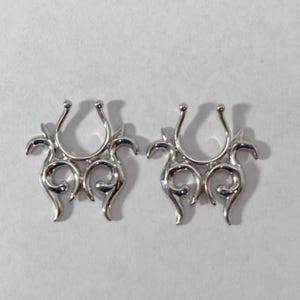 May include: Two silver nose rings with a decorative, ornate design. The rings are shaped like a horseshoe with a floral pattern.