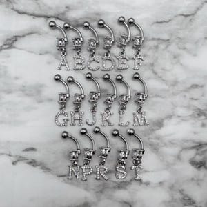 May include: A set of 16 silver belly button rings with dangling charms. Each charm is a letter of the alphabet, set with clear rhinestones.