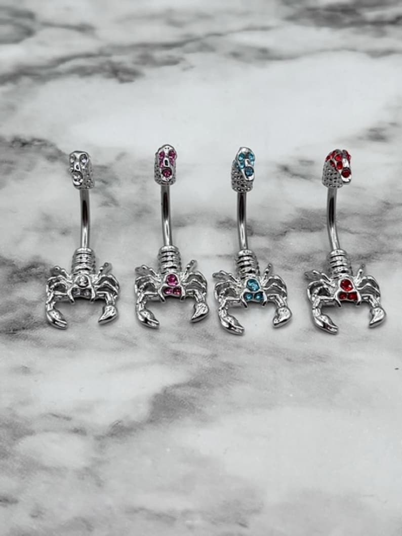 Pode incluir: Quatro an&eacute;is de umbigo de prata com pingentes de escorpi&atilde;o. Cada anel tem uma pedra preciosa de cor diferente: rosa, azul, azul claro e vermelho.
