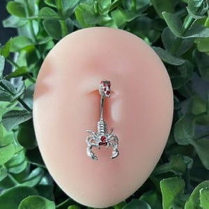 Pode incluir: Um piercing de umbigo prateado com um charme de escorpi&atilde;o cravejado de pedras vermelhas.
