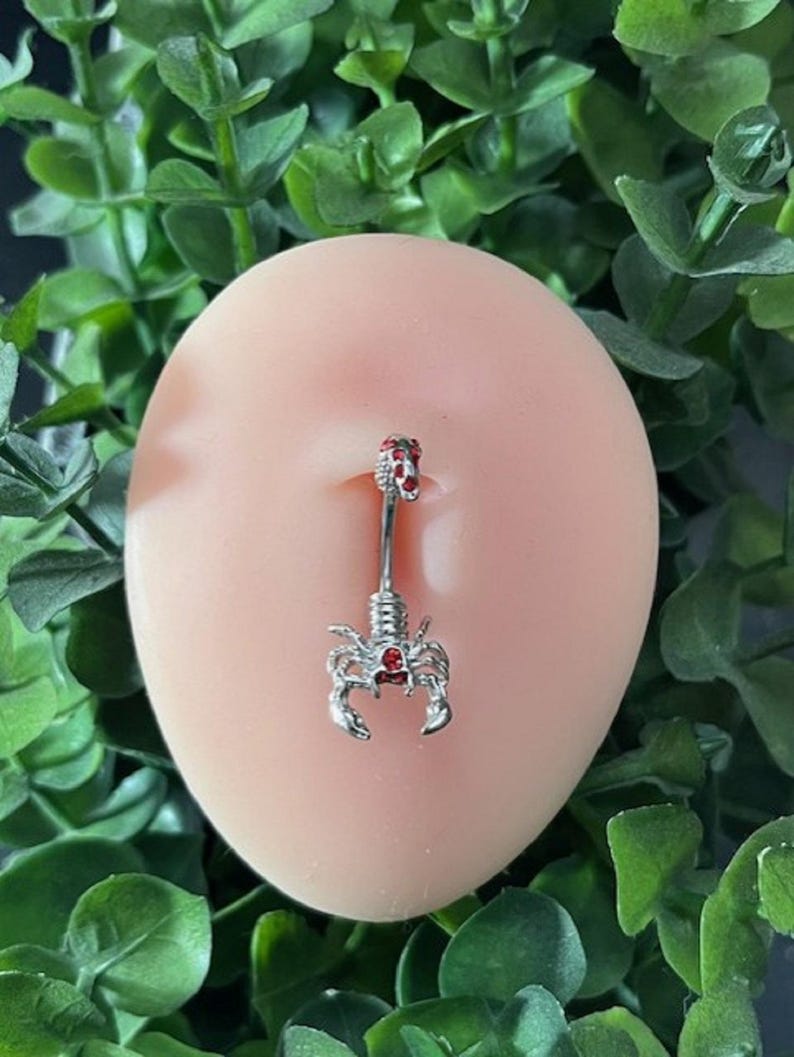 Pode incluir: Um piercing de umbigo prateado com um charme de escorpi&atilde;o cravejado de pedras vermelhas.