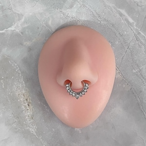 May include: A silver-toned septum ring featuring a semi-circular design adorned with a row of small, clear, round-cut stones. The jewelry is displayed on a smooth, flesh-toned anatomical model of a nose, showcasing the placement of the piece.