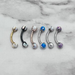 May include: Six curved barbell body jewelry pieces with opal-like gemstones. The barbells are in different colors: gold, black, rose gold, silver, and purple.